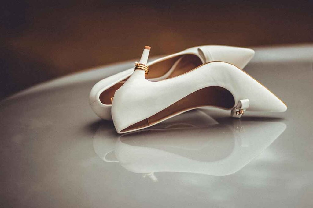 Weiße Brautschuhe mit Eheringen auf den Absätzen, elegant auf einer reflektierenden Oberfläche platziert.